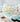 Light blue/turquoise cake with white/cream lambeth icing (very fancy) all over and florets of icing on top in the centre. There is a cup of tea in a fancy floral purple teacup with a saucer on the left. On the right in the foreground there is chocolate cake with thick blue buttercream frosting and thick blue and white lambeth icing on top. There is a single white flower in a glass vase with a cork stopper in the background as well as more floral teacups and a periwinkle blue tea pot.