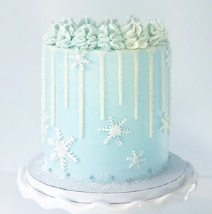 Cake covered in light blue/turquoise buttercream with white to light blue rosettes on top, white buttercream icing dripping down the sides and candy/sugar snowflake accents of varying sizes all over the mid-base of the cake. Some frost sugar effect at the complete base with little edible tiny marbles at the base. Sitting on a cake board on top of a white ceramic wavy cake stand. 