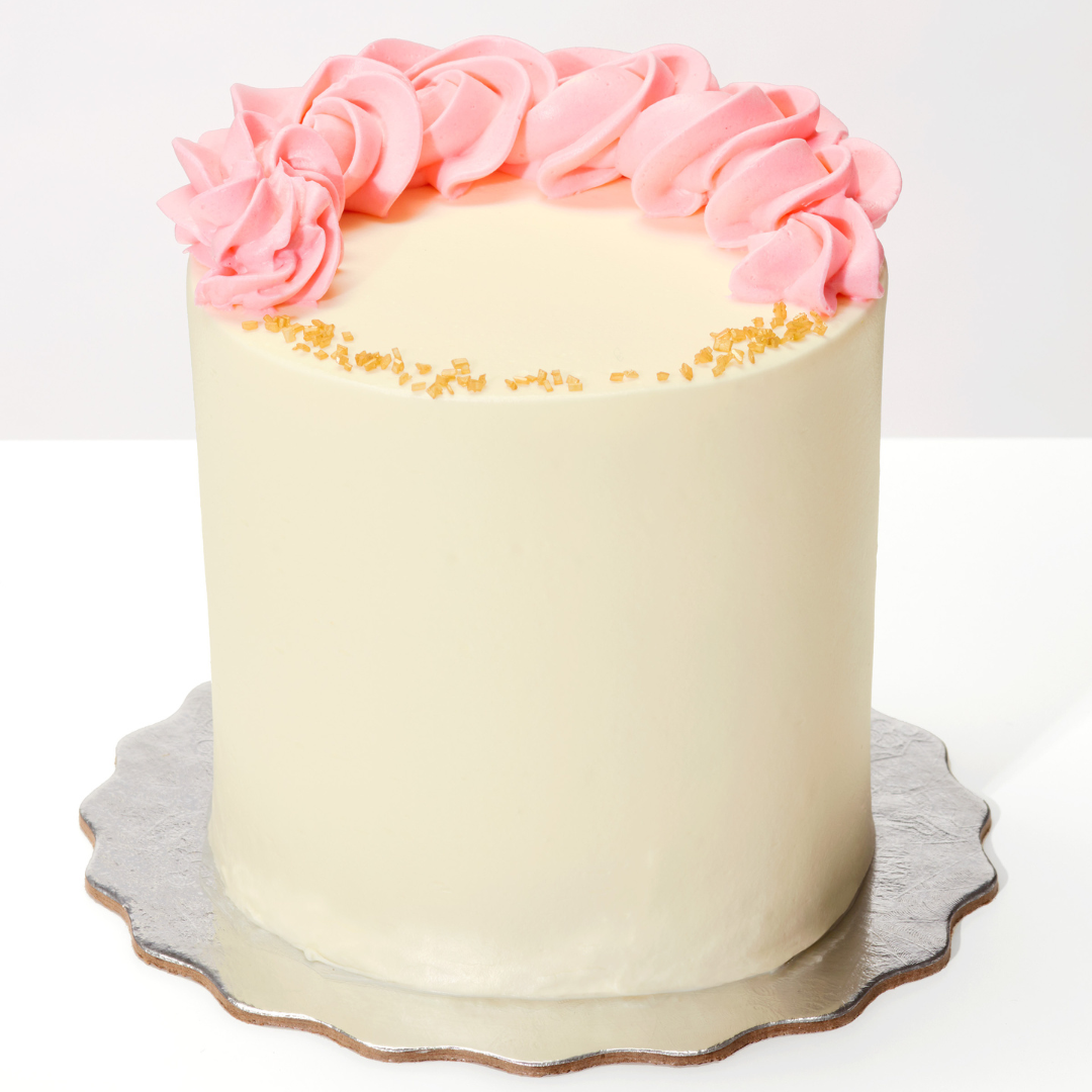Tall cake with vanilla buttercream frosting on a silver cake board, it has thick pink buttercream flowers on top in a half moon/crescent and little yellow sugar rocks on the part of the cake that has no flowers.