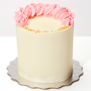 Tall cake with vanilla buttercream frosting on a silver cake board, it has thick pink buttercream flowers on top in a half moon/crescent and little yellow sugar rocks on the part of the cake that has no flowers.