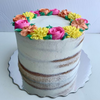 3 tier "naked" style cake with buttercream frosting all over it with some parts of the cake showing, beautiful multicoloured buttercream flowers are on top of the cake. Pink roses, yellow chrysanthemums, and orange lilies with green leaves in between where the flowers decorate the cake like a crown. Looks delicious. 